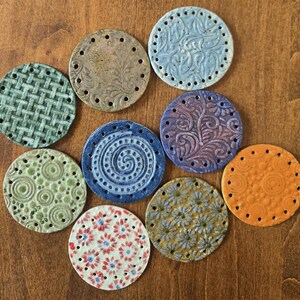 May include: Nine circular ceramic discs with various embossed patterns and perimeter holes. The collection includes textures like woven grids, floral swirls, spirals, and small blossoms in colors of green, blue, brown, orange, and white with red accents.
