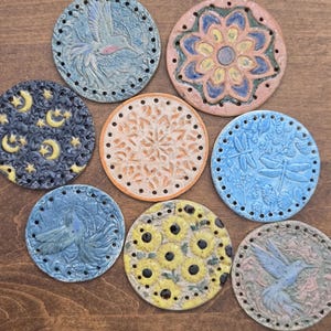 May include: Eight circular ceramic discs with pre-drilled holes along the edges. Designs include a hummingbird, a floral mandala, yellow moons and stars on black, a geometric pattern, dragonflies, a bird, and yellow sunflowers on a cream base.