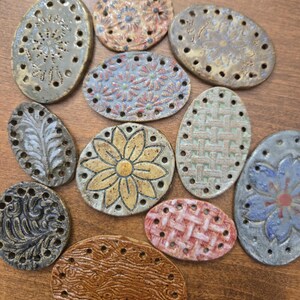 May include: A collection of eleven oval-shaped ceramic pieces, each featuring a unique embossed pattern such as flowers, leaves, or woven textures. These flat, multi-colored clay items are perforated with small holes along their outer edges.