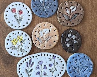 May include: A collection of eight ceramic discs and one oval piece, each featuring hand-painted floral designs and a series of small holes along the perimeter. The pieces vary in colour, including white, blue, brown, and black, with painted flowers.