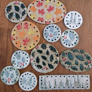 May include: A collection of ceramic craft bases in various shapes including circles, ovals, and a rectangle. Each piece features pre-drilled holes along the edges and is decorated with painted motifs such as autumn leaves, holly, and pine trees.