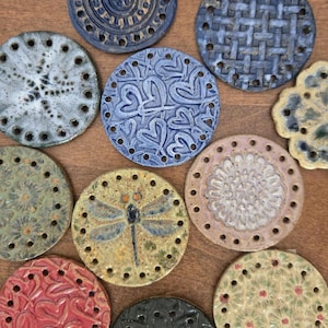 Pode incluir: Uma coleção de discos cerâmicos circulares com vários padrões em relevo, incluindo uma libélula, corações, flores e tramas geométricas. Cada disco tem uma série de pequenos orifícios perfurados ao redor do perímetro.