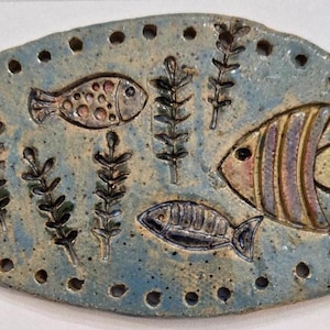 Könnte beinhalten: Eine ovale, blaue Keramikplatte mit einer geprägten Unterwasserszene, die drei verschiedene Fische und mehrere Wasserpflanzen zeigt. Der Rand der Platte ist mit einer Reihe kleiner, gleichmäßig verteilter kreisförmiger Löcher versehen.