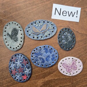May include: Six oval ceramic plaques with pre-drilled holes along the edges. Designs include a butterfly, two birds on a branch, a leafy stem, red and white stars, multiple butterflies, and a single butterfly. A white sign reads New! in black text.
