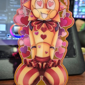 Może przedstawiać: Żółta postać słońca z czerwonymi sercami jako oczami, ubrana w czerwoną i białą spódnicę w paski i czerwoną kokardkę. Postać jest otoczona różowymi sercami i stoi na zielonej podstawie.