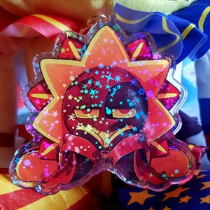 May include: An iridescent acrylic charm of a cartoon sun character with a grumpy expression. The charm is covered in glitter and stars, and is red and orange in colour.