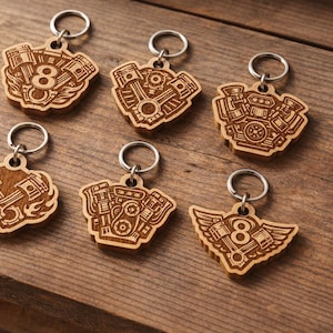 May include: Six wooden keychains with engine designs and silver key rings. The keychains feature various engine styles, including V8 engines, pistons, and winged designs. The keychains are made of light brown wood and are displayed on a wooden surface.
