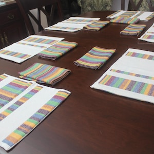 May include: A collection of white placemats and matching napkins with colorful striped accents. The stripes feature a mix of purple, green, yellow, and orange. The items are arranged on a dark brown wooden table.