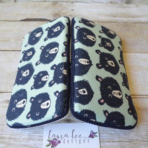May include: Two matching eyeglass cases with a light green background and a pattern of dark blue bear faces. The cases are trimmed with a dark blue rope-like border. The cases are on a light brown wooden surface.