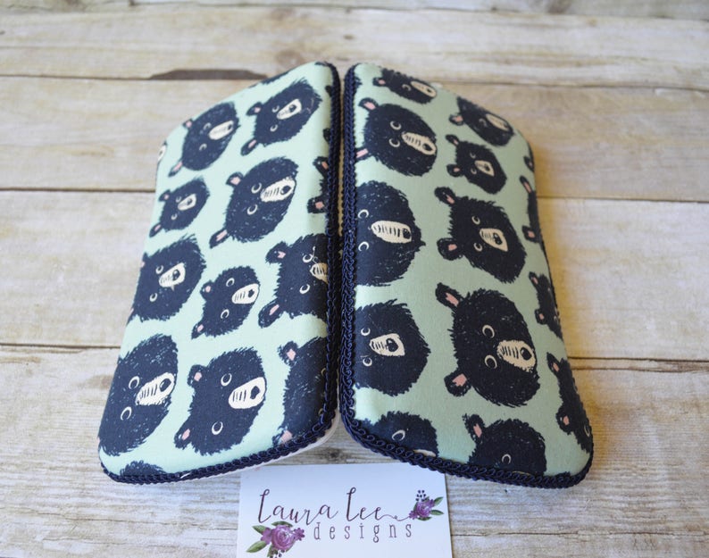 May include: Two matching eyeglass cases with a light green background and a pattern of dark blue bear faces. The cases are trimmed with a dark blue rope-like border. The cases are on a light brown wooden surface.