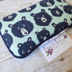 May include: A rectangular, padded case with a light teal background and a repeating pattern of black bear faces. The case is trimmed with a dark blue border and a white label with the text "Laura Lee Designs".