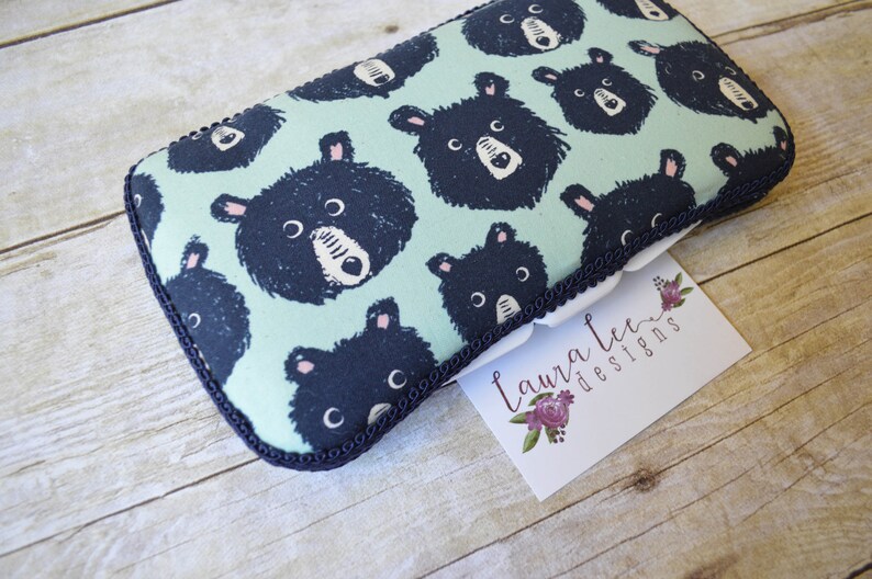 May include: A rectangular, padded case with a light teal background and a repeating pattern of black bear faces. The case is trimmed with a dark blue border and a white label with the text "Laura Lee Designs".