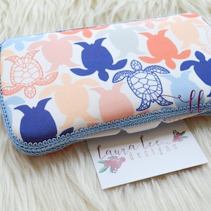 May include: A blue and white fabric tissue box cover with a sea turtle pattern. The cover has a blue trim and a white label with the text "Laura Lee Designs".