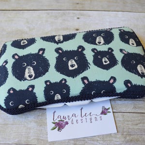 May include: Rectangular fabric pouch with a repeating pattern of black bear faces on a light teal background. The pouch is trimmed with a dark navy blue border and a decorative black trim. A small white card with the text "Laura Lee Designs" is in the foreground.