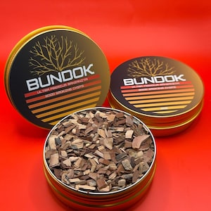 May include: Two gold tins of wood smoking chips labelled "Bundok Ultra Premium Manzanita Wood Smoking Chips". One tin is open and shows the wood chips inside.
