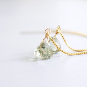 Może przedstawiać: Para delikatnych złotych kolczyków z łańcuszkiem i zielonymi kamieniami. Kolczyki są połączone długim łańcuszkiem.