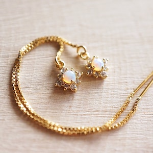 May include: Gold-coloured earrings with a floral design. Each earring has a central opal-like stone surrounded by small, clear crystals. The earrings are connected by a delicate chain, creating a stylish and elegant look. The background is a light wooden surface.