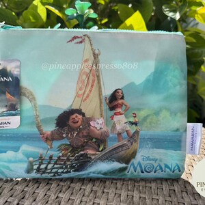 Puede incluir: Bolsa de cosm&eacute;ticos azul claro con tem&aacute;tica de Disney Moana con personajes en un barco. La bolsa tiene cierre de cremallera y una etiqueta con el logotipo de Disney Moana y la marca Hawaiian Airlines. El fondo muestra una escena oce&aacute;nica tropical.