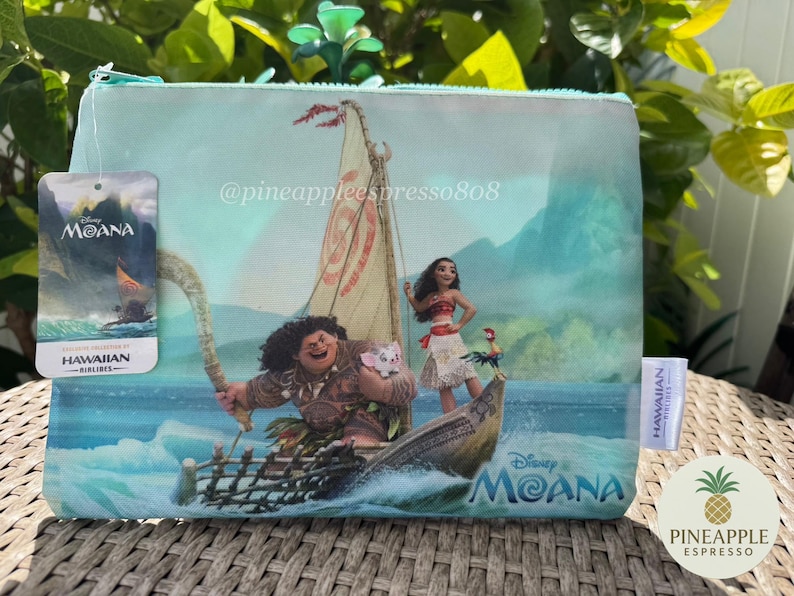 Puede incluir: Bolsa de cosm&eacute;ticos azul claro con tem&aacute;tica de Disney Moana con personajes en un barco. La bolsa tiene cierre de cremallera y una etiqueta con el logotipo de Disney Moana y la marca Hawaiian Airlines. El fondo muestra una escena oce&aacute;nica tropical.