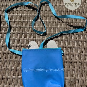 Puede incluir: Un peque&ntilde;o bolso bandolera azul brillante con una correa a juego. El bolso tiene cierre de cremallera y contiene dos objetos peque&ntilde;os de color claro. El texto "@pineappleespresso808" est&aacute; impreso en el bolso. Un logotipo de pi&ntilde;a es visible en la esquina superior derecha.