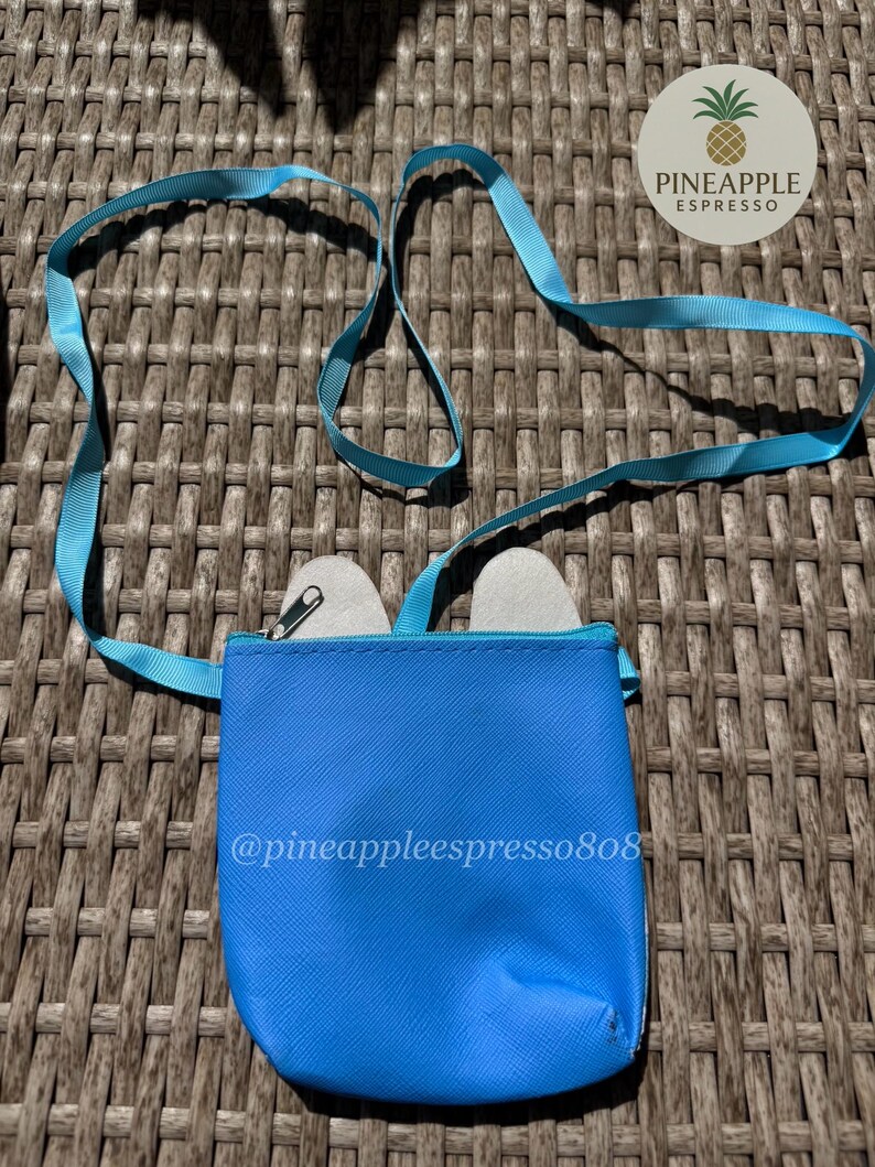 Puede incluir: Un peque&ntilde;o bolso bandolera azul brillante con una correa a juego. El bolso tiene cierre de cremallera y contiene dos objetos peque&ntilde;os de color claro. El texto "@pineappleespresso808" est&aacute; impreso en el bolso. Un logotipo de pi&ntilde;a es visible en la esquina superior derecha.