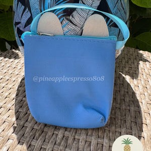 Puede incluir: Una bolsa azul claro con cremallera y asa a juego, que contiene dos objetos blancos. El texto "@pineappleespresso808" est&aacute; impreso en la bolsa. Un logotipo de pi&ntilde;a es visible en la esquina inferior derecha. La bolsa est&aacute; sobre una superficie tejida.