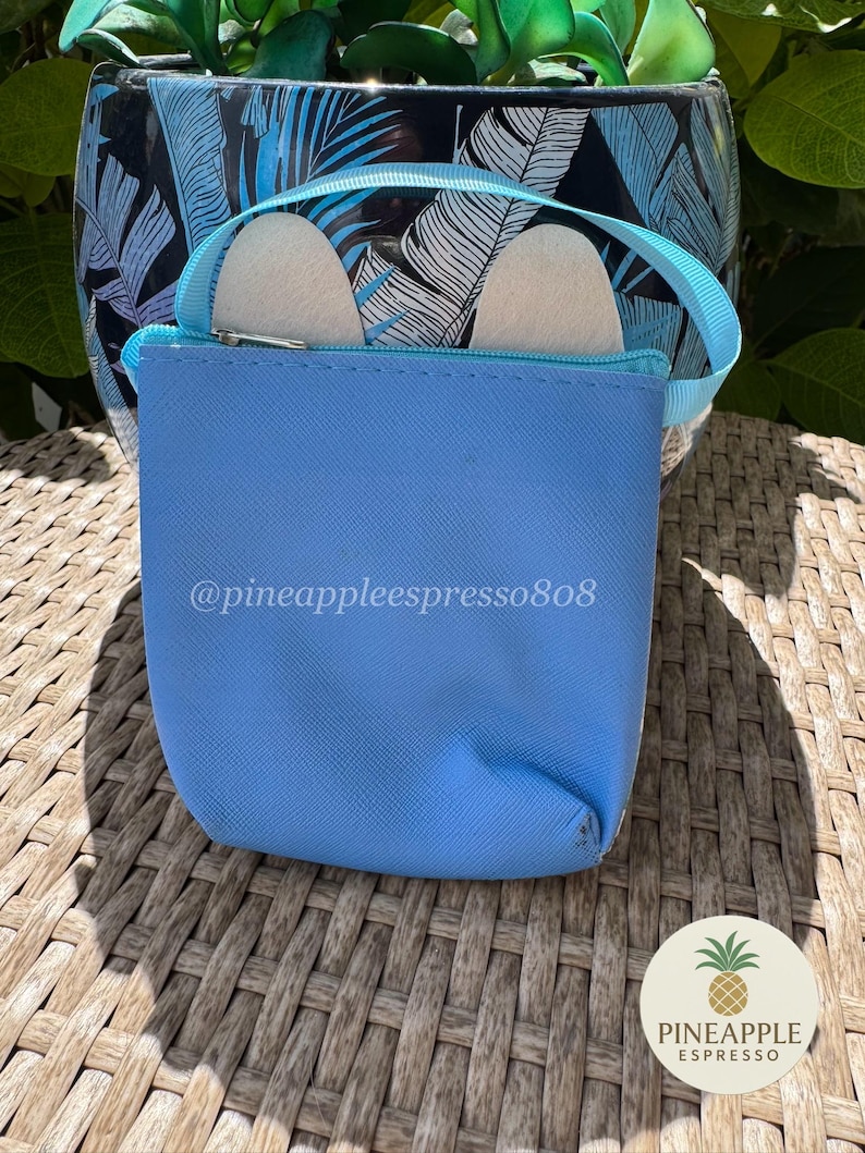 Puede incluir: Una bolsa azul claro con cremallera y asa a juego, que contiene dos objetos blancos. El texto "@pineappleespresso808" est&aacute; impreso en la bolsa. Un logotipo de pi&ntilde;a es visible en la esquina inferior derecha. La bolsa est&aacute; sobre una superficie tejida.
