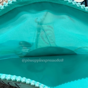 Puede incluir: Vista abierta de una bolsa con cremallera turquesa con interior turquesa. La cremallera es blanca. El texto "@pineappleespresso808" es visible. Un logotipo de Pineapple Espresso est&aacute; presente.