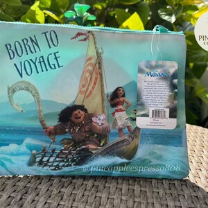 Puede incluir: Una bolsa con cremallera azul claro con una escena de la pel&iacute;cula de Disney Moana. La imagen muestra a Moana, Maui y un barco en el oc&eacute;ano, con el texto "BORN TO VOYAGE". Una etiqueta con el logotipo de Moana est&aacute; adjunta.