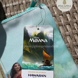 Puede incluir: Una bolsa con cremallera azul claro con un dise&ntilde;o de Disney Moana. La bolsa tiene una etiqueta que dice "Moana" y "Exclusive Collection by Hawaiian Airlines". La bolsa tiene una cremallera turquesa y una leng&uuml;eta a juego.