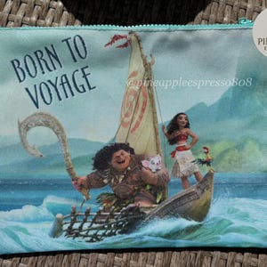 Puede incluir: Una bolsa con cremallera con una colorida ilustraci&oacute;n de personajes en un barco navegando por el oc&eacute;ano. El texto "BORN TO VOYAGE" est&aacute; impreso en la parte superior izquierda. La bolsa tiene una cremallera turquesa y una etiqueta de Hawaiian Airlines.