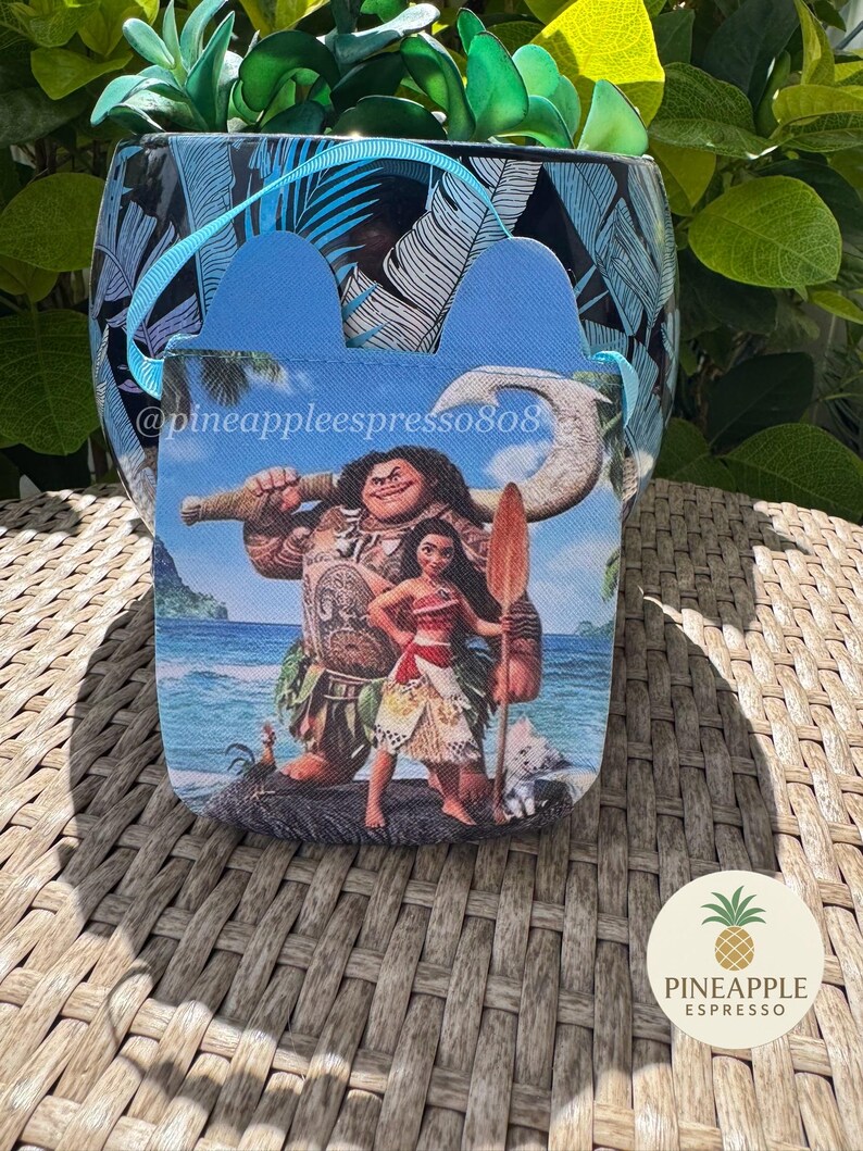 Puede incluir: Un recipiente con estampado azul y blanco con un asa azul y una peque&ntilde;a bolsa con personajes de la pel&iacute;cula Moana. La bolsa tiene un asa azul y el texto "@pineappleespresso808". Un logotipo de pi&ntilde;a es visible.