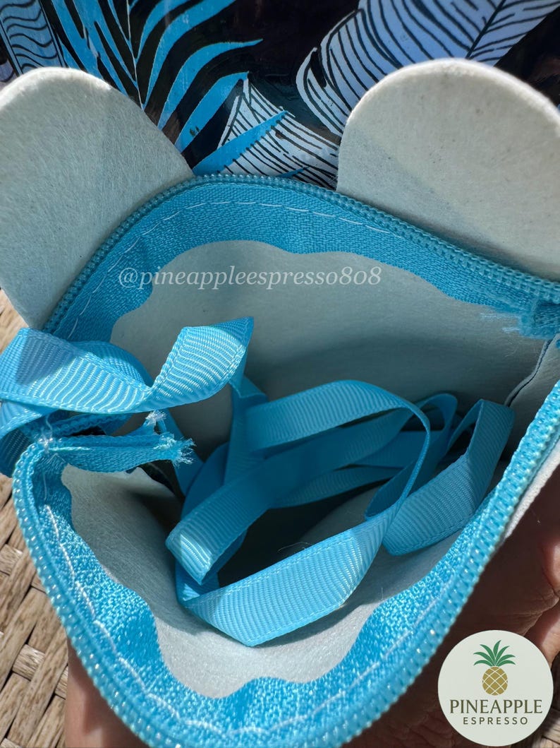 Puede incluir: Una bolsa con cremallera azul claro con detalles de fieltro blanco y una cinta azul claro. La bolsa tiene un dise&ntilde;o &uacute;nico con una cremallera azul claro y el texto "@pineappleespresso808". Se ve un logotipo de pi&ntilde;a.