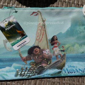 Puede incluir: Una bolsa con cremallera azul claro con un dise&ntilde;o de Disney Moana. La bolsa muestra personajes de la pel&iacute;cula en un barco, con una etiqueta que dice "Moana" y "Hawaiian Airlines". La bolsa tiene una cremallera turquesa y una peque&ntilde;a etiqueta blanca.