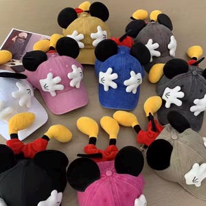 May include: A selection of baseball caps in shades of pink, blue, and yellow. Each cap is adorned with large black ears, white gloved hands, and a Mickey Mouse design. The caps are displayed on a light-toned surface.