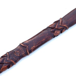 May include: Close-up of a carved wooden staff with a dark brown finish. The staff features intricate details, including a braided pattern, diamond shapes, and leaf-like accents. The design suggests a fantasy or historical theme.