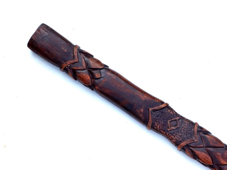 May include: A dark brown, carved wooden staff or handle. The wood has a rich, warm tone with intricate leaf and diamond patterns carved along its length. The end is a solid, unadorned cylinder.