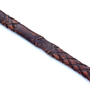 May include: A dark brown, carved wooden handle with intricate braided and geometric patterns. The handle is likely for a tool or weapon, showcasing detailed craftsmanship and a rustic aesthetic.