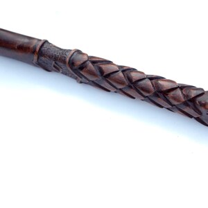 May include: Close-up of a dark brown, intricately carved wooden wand. The wand features a woven pattern along the handle and a smooth, rounded tip. The wood has a rich, warm tone, suggesting a handcrafted item.