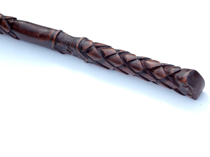 May include: Close-up of a dark brown, intricately carved wooden wand. The wand features a woven pattern along the handle and a smooth, rounded tip. The wood has a rich, warm tone, suggesting a handcrafted item.