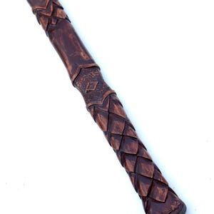 May include: A brown, carved wooden handle with a textured, scale-like pattern. The handle has a dark finish and is angled diagonally against a white background. The design includes diamond and leaf-like details.