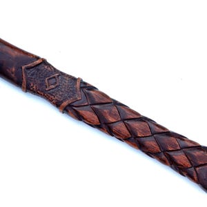 May include: Close-up of a wooden axe handle with intricate carvings. The handle features a dark brown, woven pattern and a decorative section with a diamond-shaped design. The end of the handle has a carved symbol.