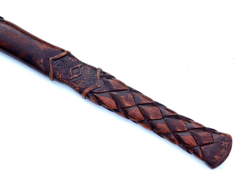 May include: Close-up of a wooden axe handle with intricate carvings. The handle features a dark brown, woven pattern and a decorative section with a diamond-shaped design. The end of the handle has a carved symbol.