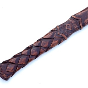 May include: Close-up of a wooden axe handle with intricate carved details. The handle features a woven pattern and a decorative end with a cross design. The wood has a rich, dark brown color.