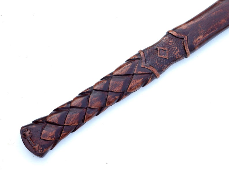 May include: Close-up of a wooden axe handle with intricate carved details. The handle features a woven pattern and a decorative end with a cross design. The wood has a rich, dark brown color.