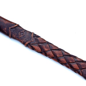 May include: Close-up of a wooden axe handle with intricate carvings. The handle features a dark brown, woven pattern and a decorative section with a diamond-shaped design. The end of the handle has a carved symbol.