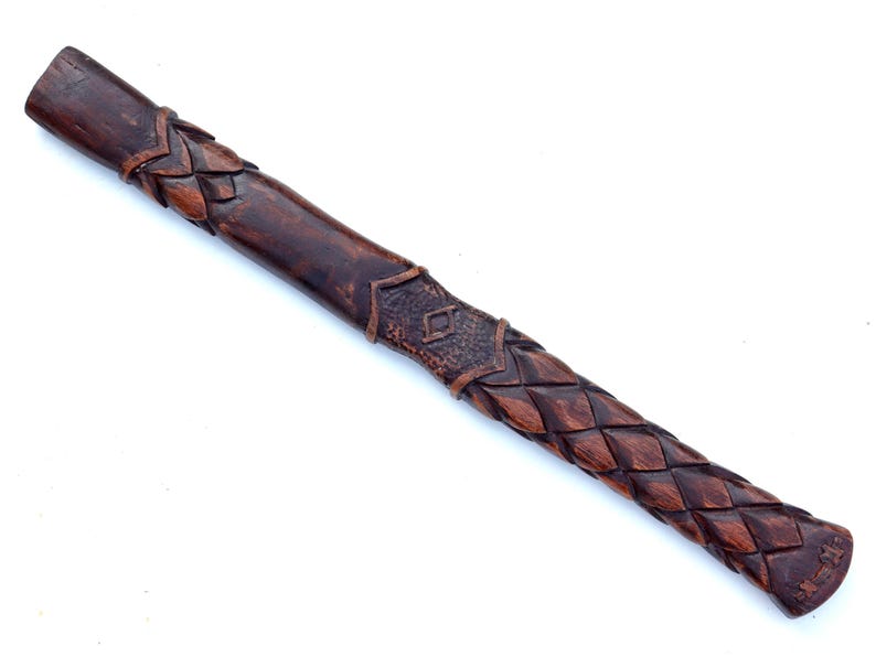 May include: A dark brown, carved wooden handle with intricate braided and geometric patterns. The handle is likely for a tool or weapon, with a textured grip and decorative elements.