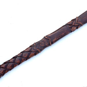 May include: A handcrafted wooden handle with intricate carved details. The handle features a dark brown finish with a braided pattern on the grip and decorative elements near the top. The design suggests a tool or weapon handle.