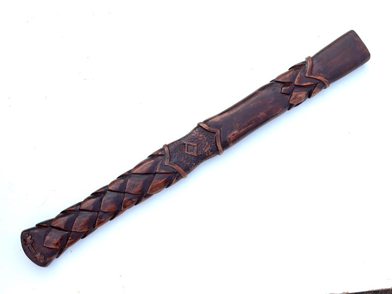 May include: A handcrafted wooden handle with intricate carved details. The handle features a dark brown finish with a braided pattern on the grip and decorative elements near the top. The design suggests a tool or weapon handle.