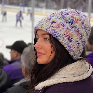 May include: A chunky knit beanie featuring a multi-coloured speckled yarn pattern with hues of purple, blue, yellow, and pink. The hat has a thick, ribbed texture and a folded brim, crafted from a soft, bulky yarn that creates a distinct, raised stitch.
